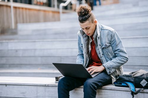 Free Concentrated male freelancer in denim jacket typing on netbook while sitting on wooden steps with backpack on street on blurred background Stock Photo