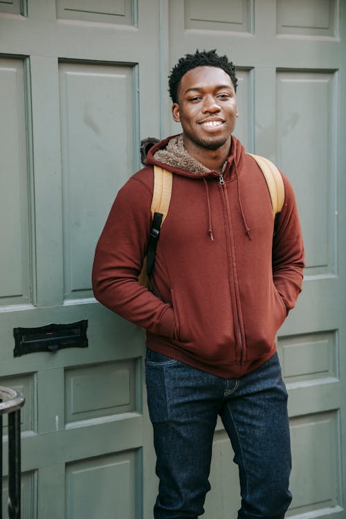 Free Smiling African American male with backpack looking at camera while standing near entrance of building with hands in pockets on street Stock Photo