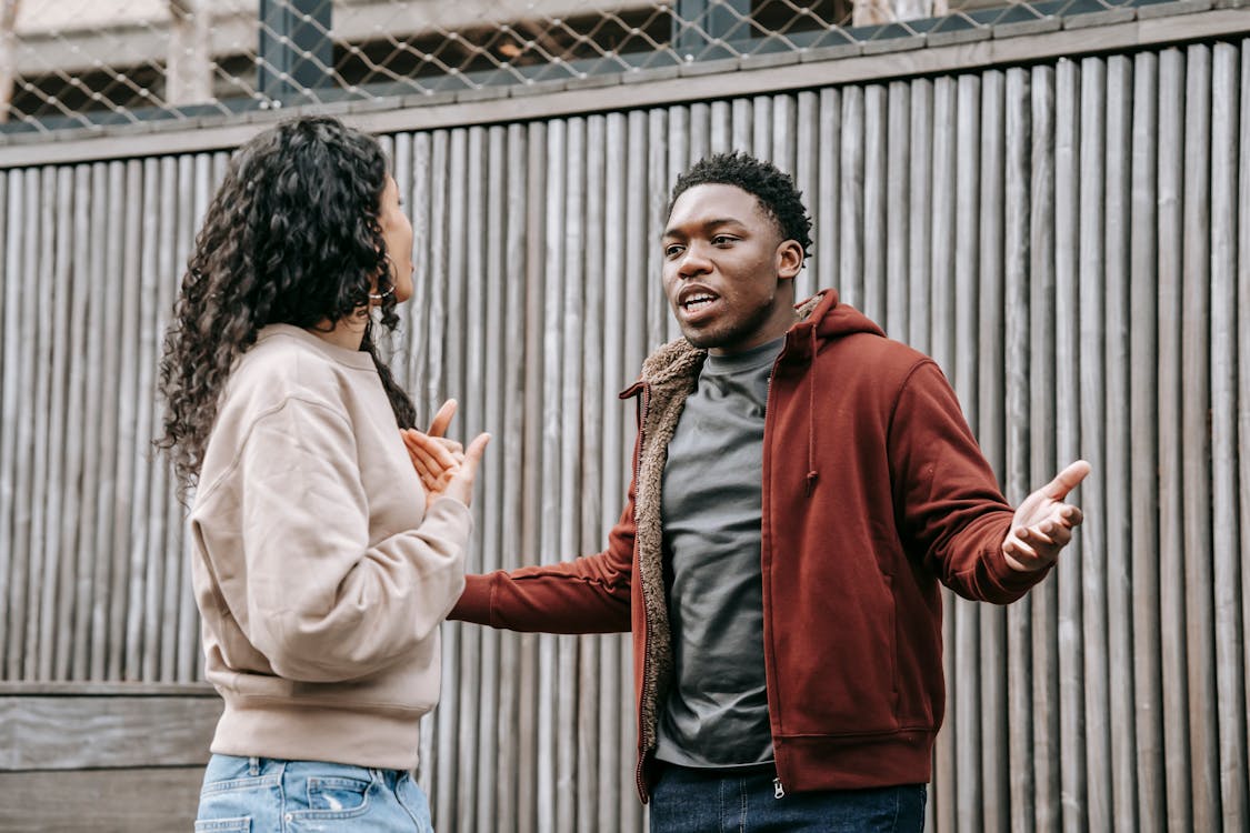 Free Angry African American boyfriend and ethnic girlfriend looking at each other while having emotional dispute on street near wooden fence Stock Photo