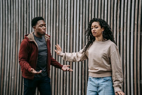 Insulted ethnic girlfriend with outstretched arm and irritated African American boyfriend having quarrel near wooden wall on street during breakup