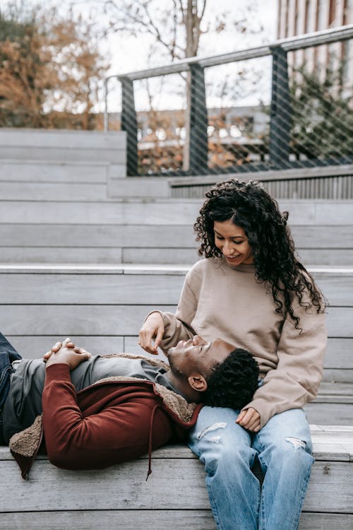 Content African American man with clasped hands lying on lap of happy ethnic woman and looking at each other while sitting on stairs in park with metal grid during date