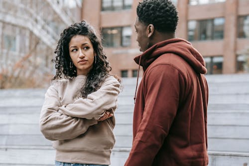 Unset ethnic woman with crossed arms and African American man looking at each other while having quarrel on street with building on blurred background