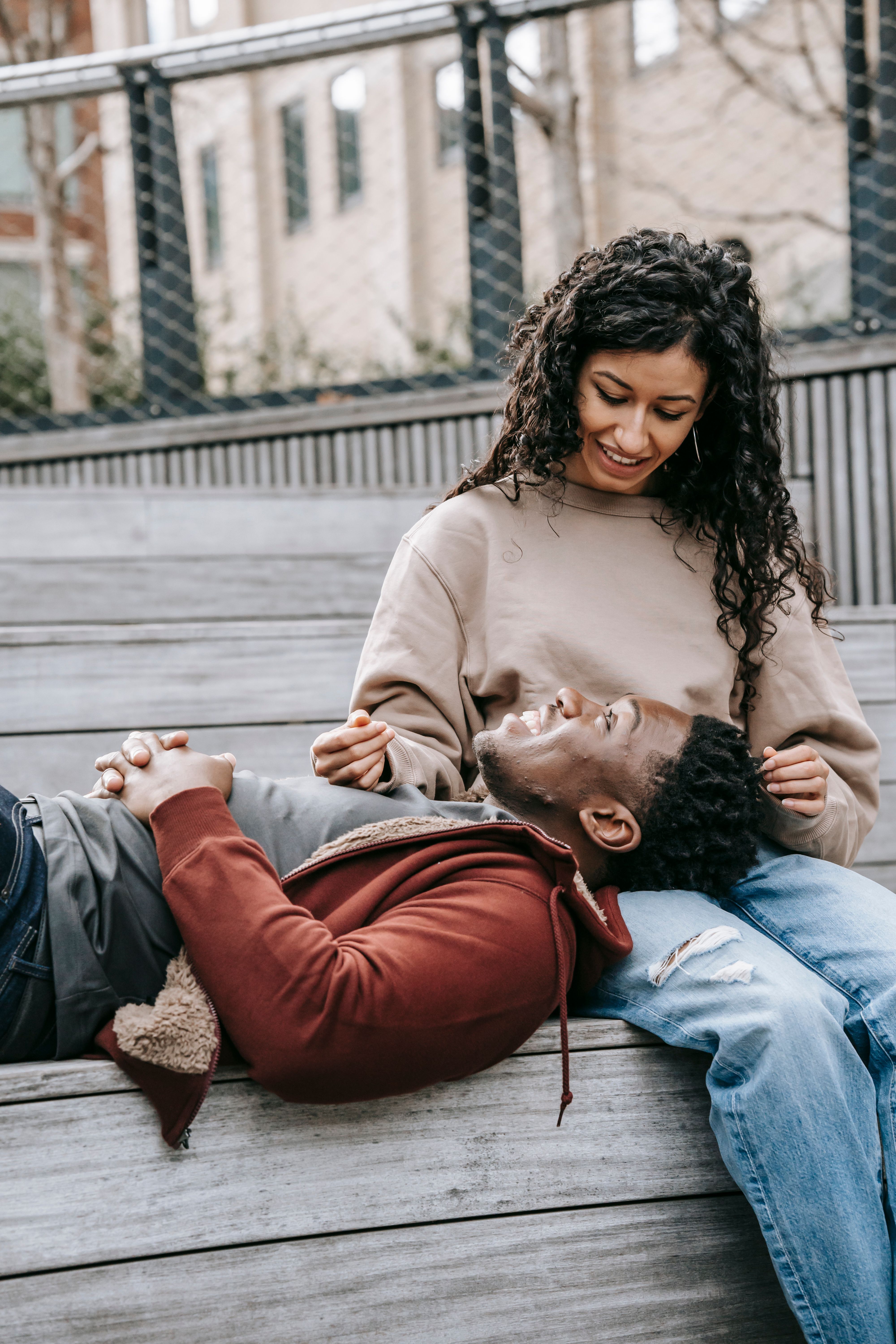 Cheerful African American boyfriend with clasped hands lying on lap of ethnic girlfriend while resting on wooden bench and looking at each other in park with metal grid