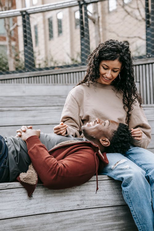 Cheerful African American boyfriend with clasped hands lying on lap of ethnic girlfriend while resting on wooden bench and looking at each other in park with metal grid