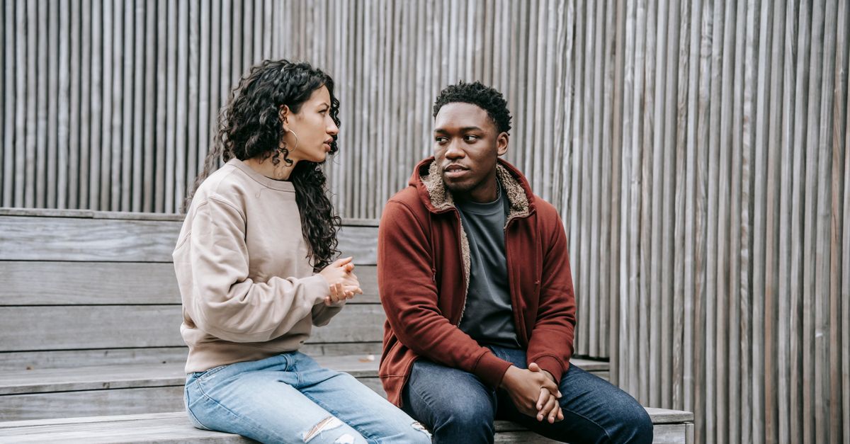 Frustrated ethnic girlfriend and African American boyfriend sitting on wooden step and looking at each other while having dispute on street