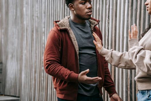 Unrecognizable woman with outstretched arm trying to stop irritated African American man while having argument on street near wooden fence