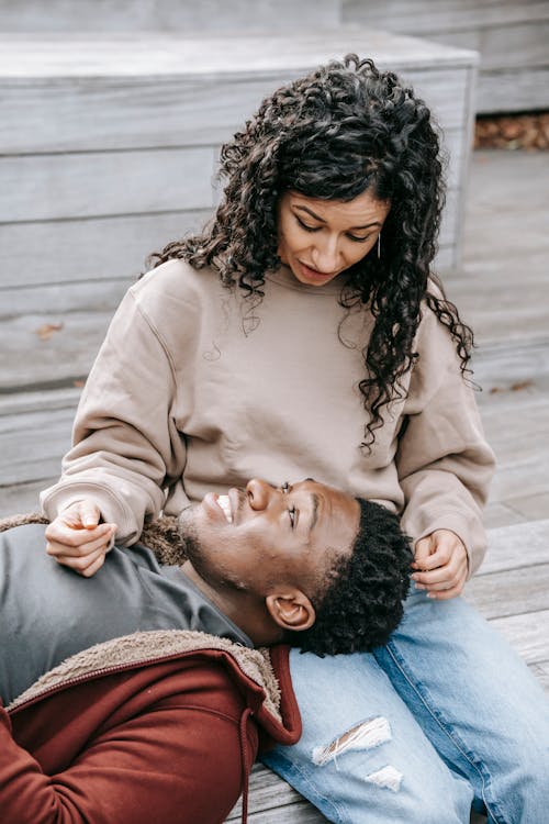 Cheerful African American boyfriend lying on lap of ethnic girlfriend while sitting on wooden steps on street and looking at each other