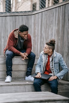 Two young men chatting on wooden steps, capturing a casual and friendly interaction in daylight.
