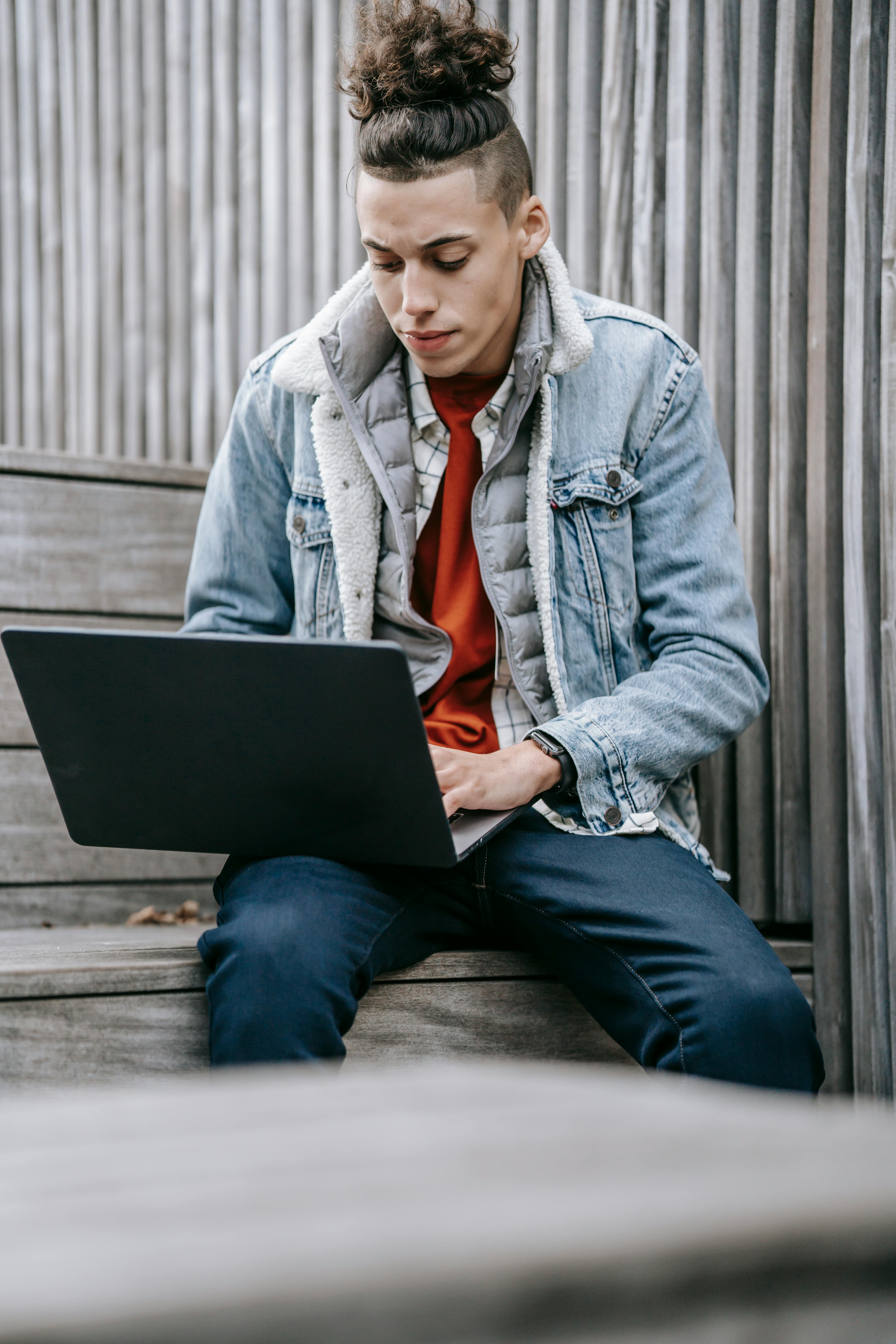 Stylish young man using a laptop on wooden steps outdoors, representing urban digital lifestyle.