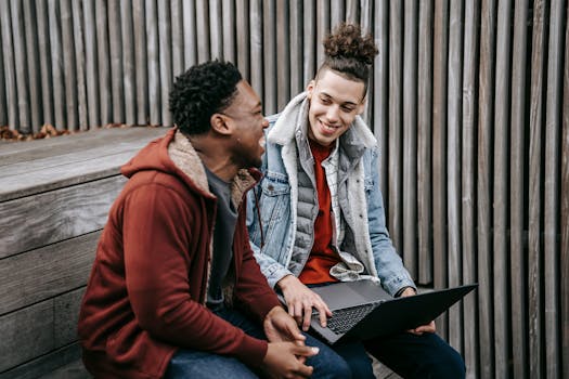 Cheerful multiracial students talking and smiling while surfing internet on netbook on wooden stairs near fence in daytime