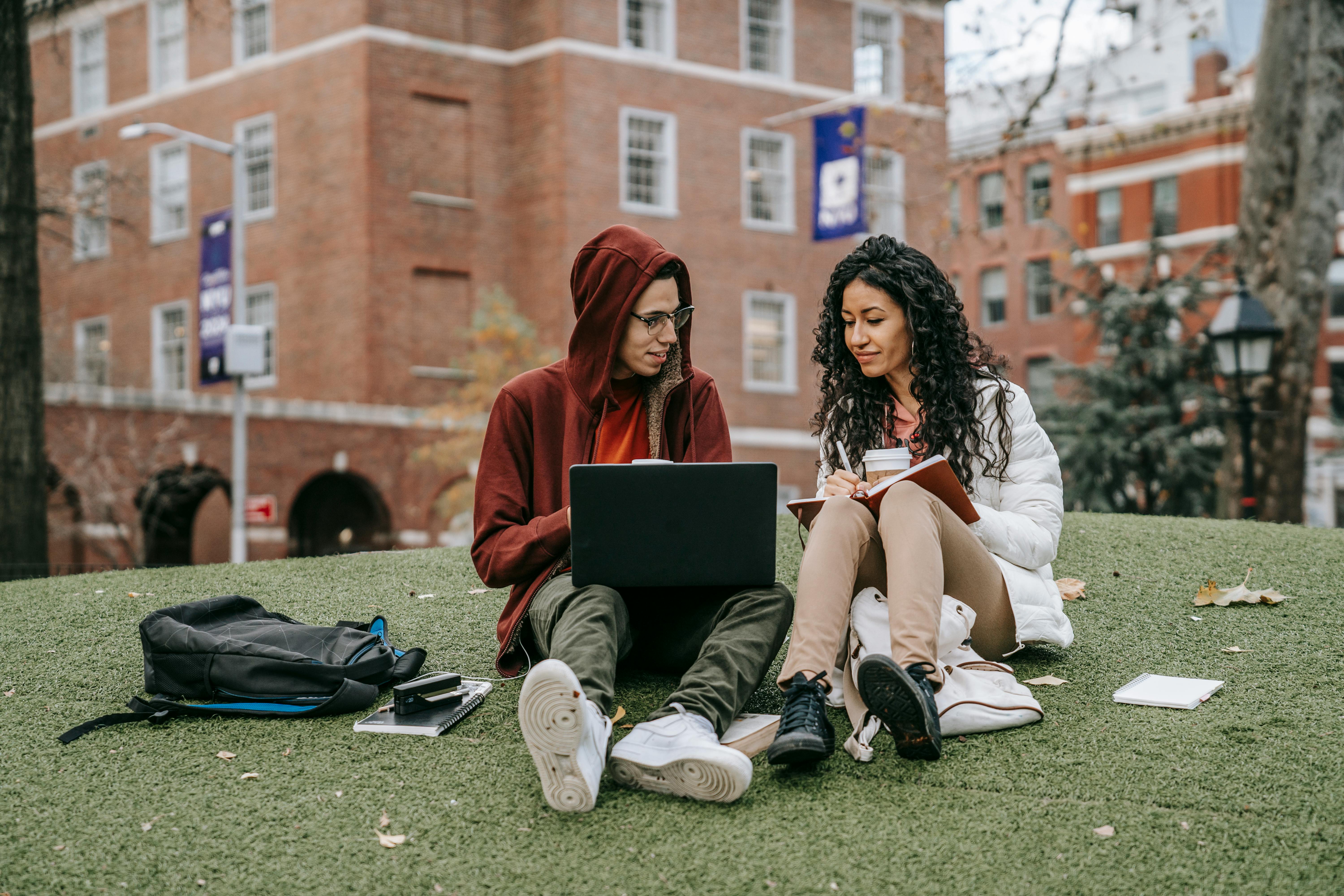 Two college students study together on campus lawn with laptop and notes.