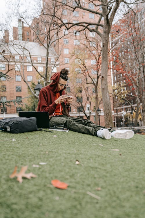 Low angle of young student in casual wear and eyeglasses sitting on lawn in park with laptop and browsing mobile phone