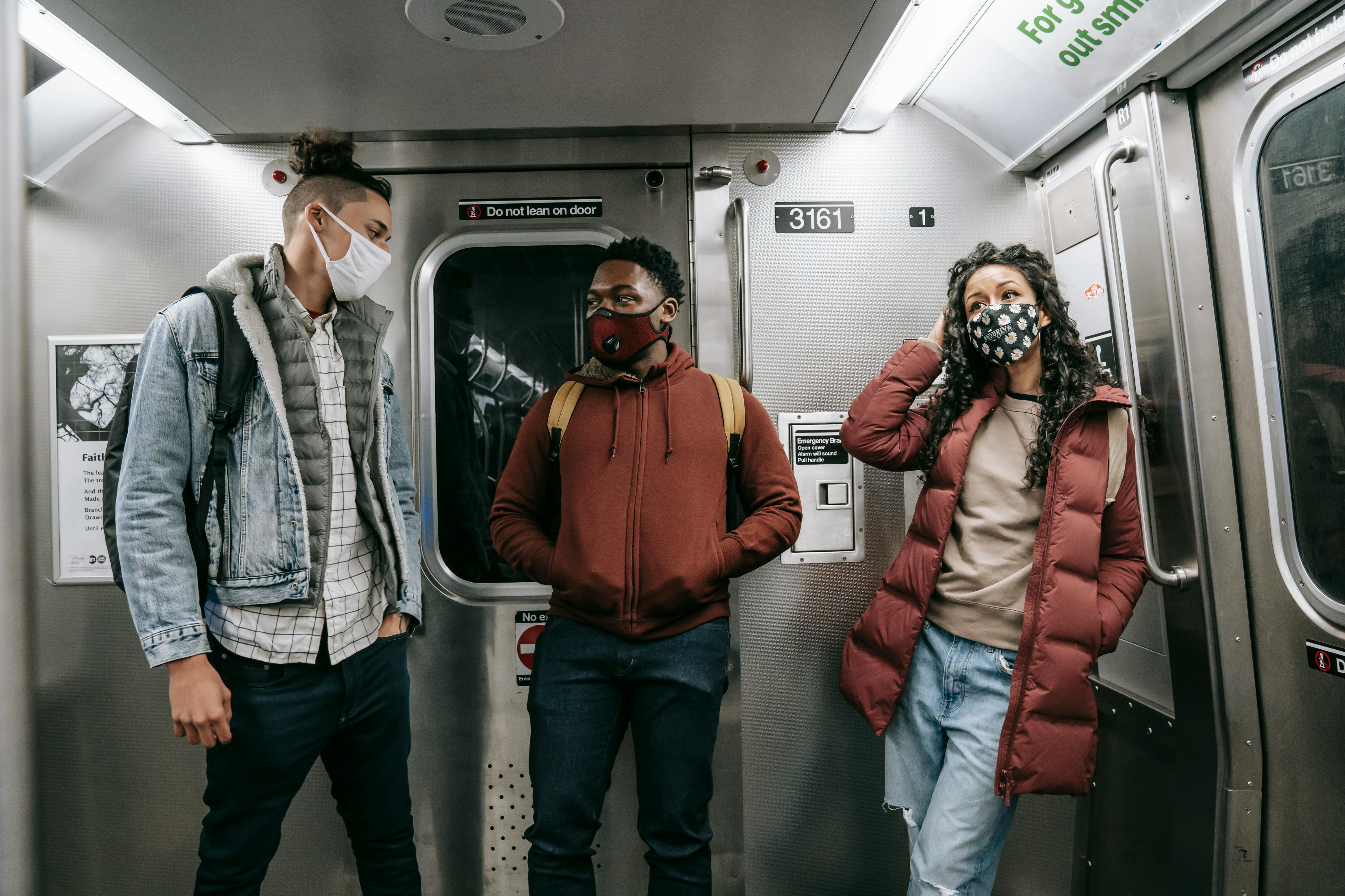 Group of faceless young multiracial people in warm clothes and protective masks wearing backpacks while standing in light train in metro