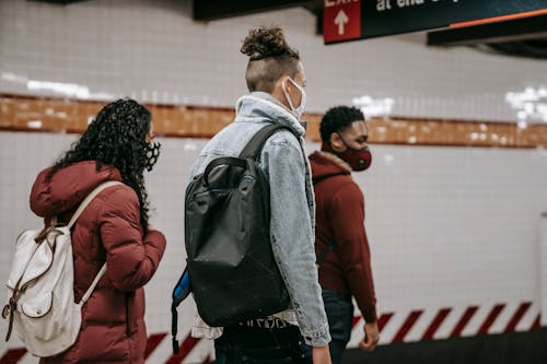 Free Side view of anonymous young multiracial friends in warm clothes and protective masks with backpacks walking on underground station Stock Photo
