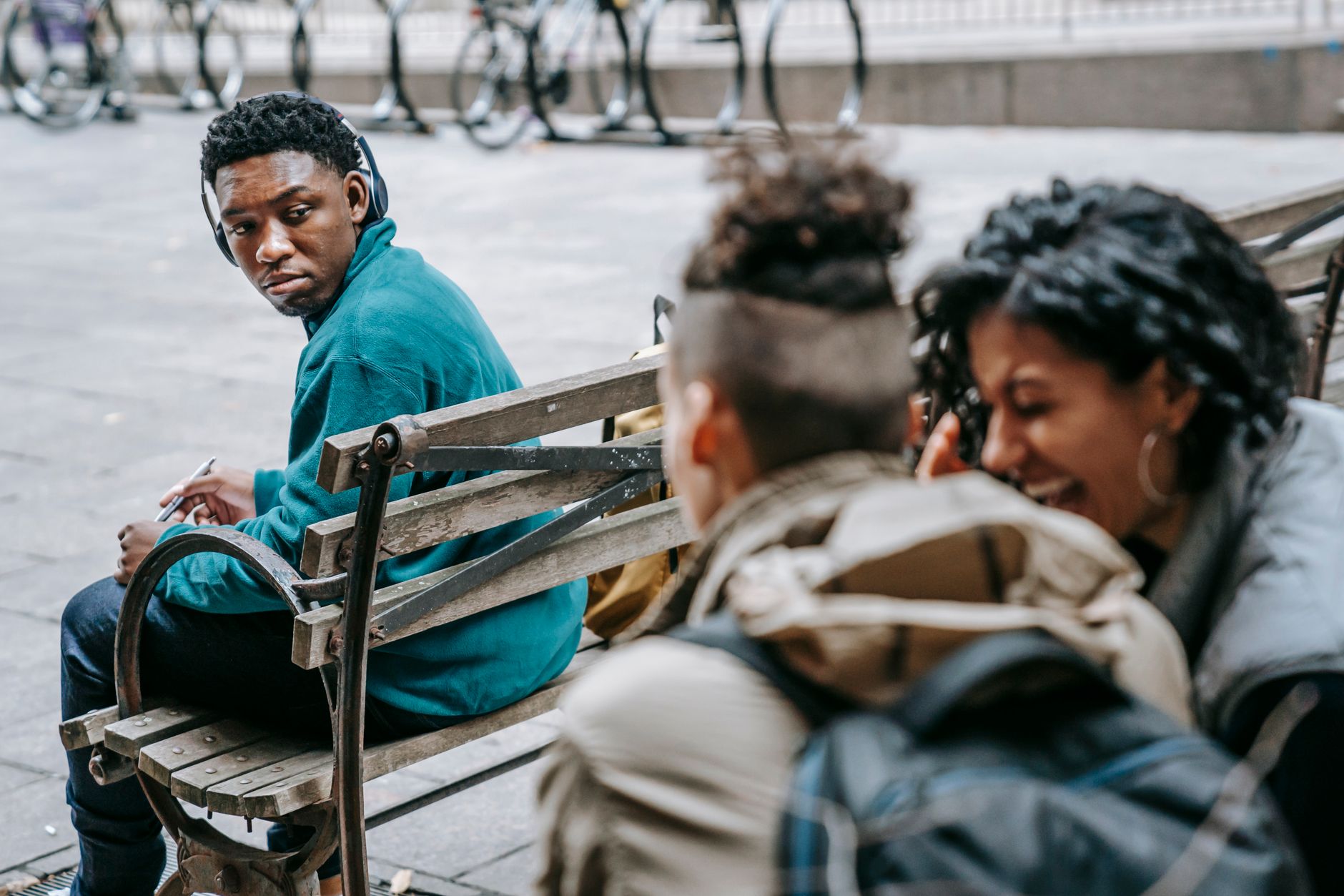 Back view of young friends in casual outfit with backpacks laughing and gossiping behind back of African American male sitting on bench in headphones in city street in daylight