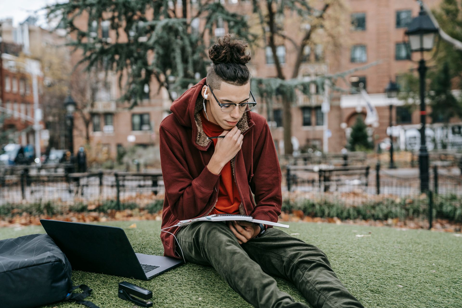 Young male student in casual outfit and earphones sitting on grassy lawn while preparing for exams with notebook and pen while using netbook near backpack and stapler in campus