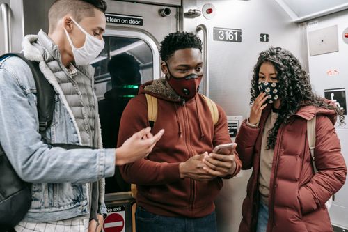 Free Group of anonymous young multiracial people in protective masks and warm clothes with backpacks talking while standing with cellphone in light train in underground Stock Photo