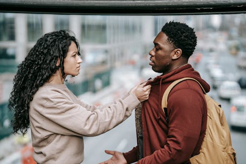Side view of young multiracial couple in casual outfit standing in city street and having argument with each other while woman holding man by jacket and looking at each other