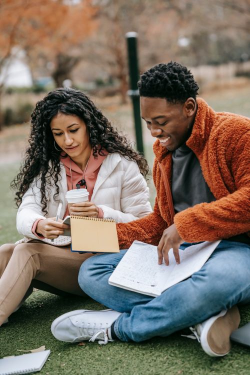 Free Young multiracial students in casual outfit and outerwear sitting on grass in park and studying with notepads and paper cup of drink in daylight Stock Photo
