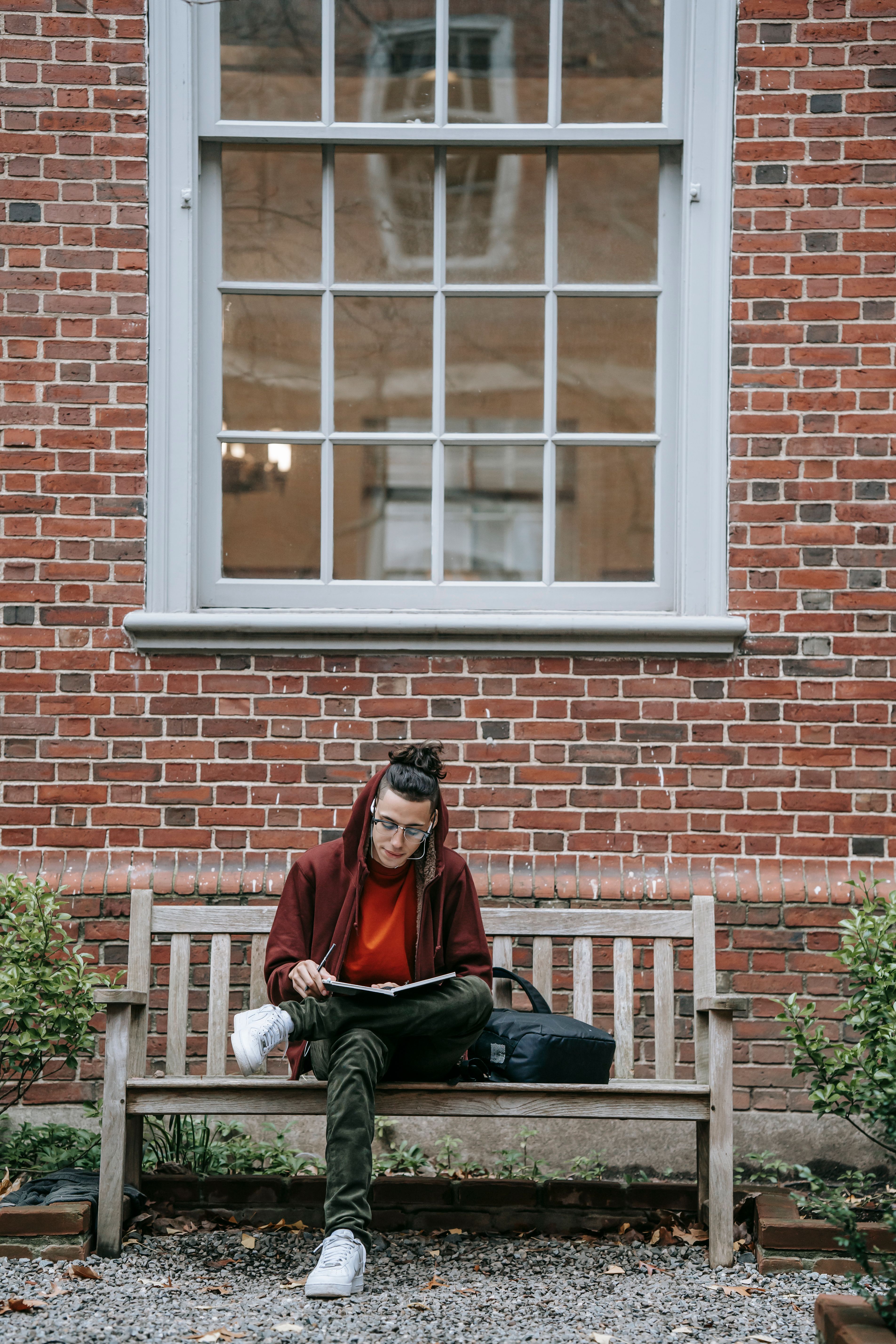 Full body of concentrated young male in casual clothes sitting on bench near building with brick wall and window while studying with notebook in campus near plants in daytime