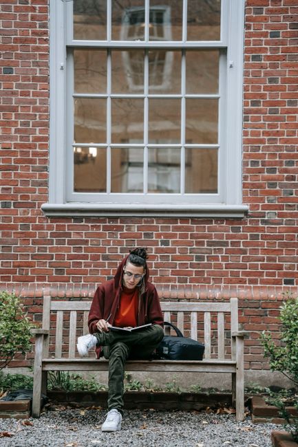 Full body of concentrated young male in casual clothes sitting on bench near building with brick wall and window while studying with notebook in campus near plants in daytime Full body of concentrated young male in casual clothes sitting on bench near building with brick wall and window while studying with notebook in campus near plants in daytime