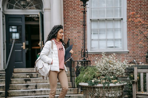 Confident young ethnic woman in warm clothes with backpack walking in city street with folder in daytime near building door and stairs near plant