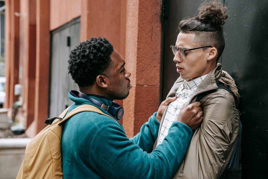 Free Side view of young African American guy in casual outfit bullying classmate by holding jacket and leaning on building wall while standing in city street with backpack in daylight Stock Photo