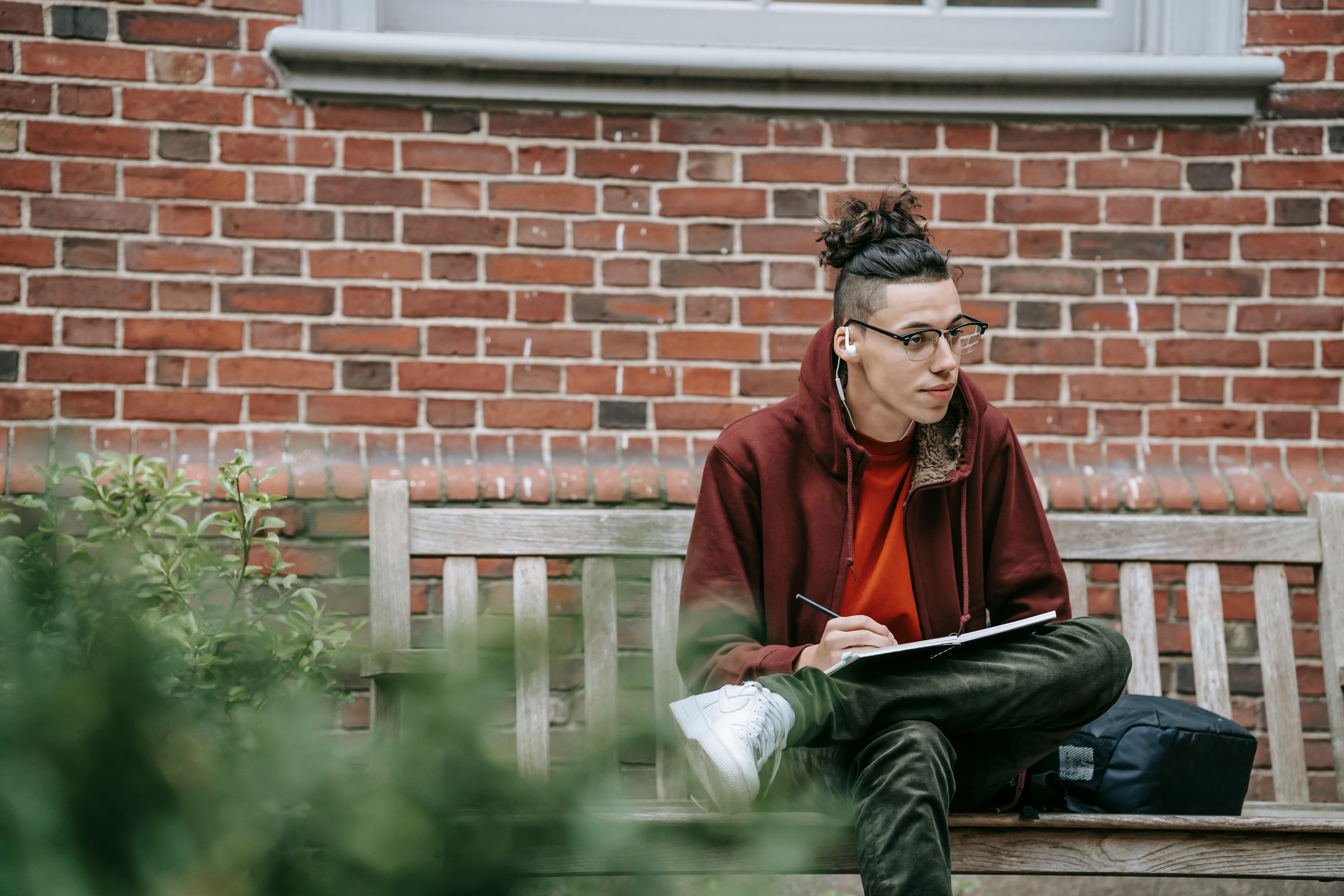 Thoughtful young guy in casual clothes with earphones sitting on bench near backpack and building while studying and taking notes in notepad with pen in campus near plants in daytime