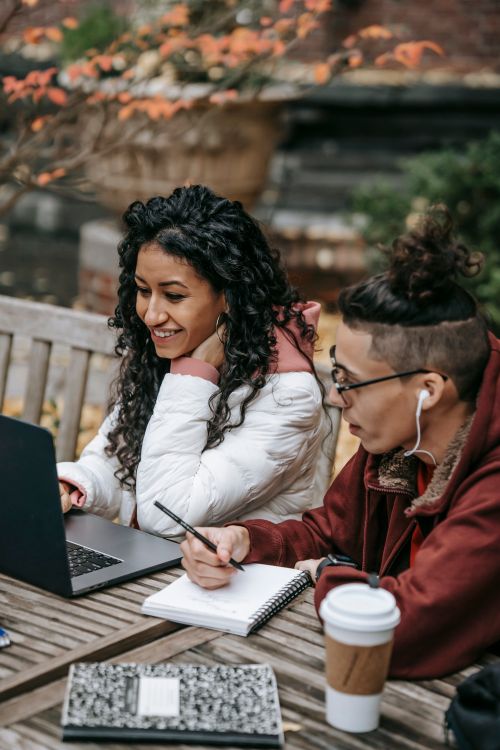 Free Young multiethnic friends in warm clothes sitting at table in campus and preparing for exam with notepad and pen while browsing on laptop near cup of drink in daytime Stock Photo