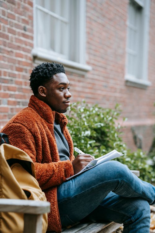 Free Side view of African American young man in casual clothes near backpack sitting on bench near building while preparing for exam while taking notes in document with pen in campus Stock Photo