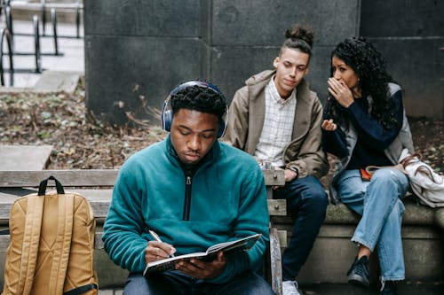 Free Young multiethnic friends in casual outfit with backpack gossiping behind back of African American guy sitting on bench in headphones and taking notes in notebook in city street in daylight Stock Photo