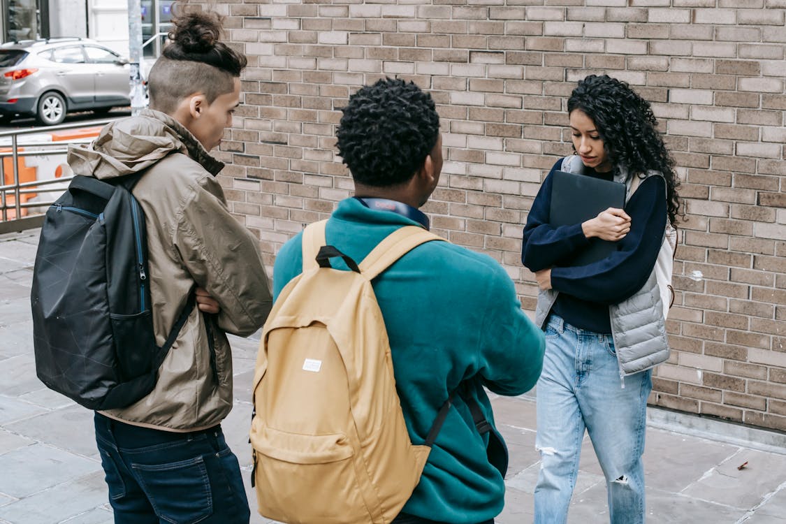 Free Young multiracial male friends with backpacks offending female with folder on city street near brick wall in daylight Stock Photo