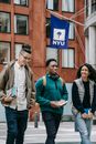 Smiling young multiracial students in casual clothes with backpacks talking while strolling in New York City with books and headphones near university building with flag in daytime