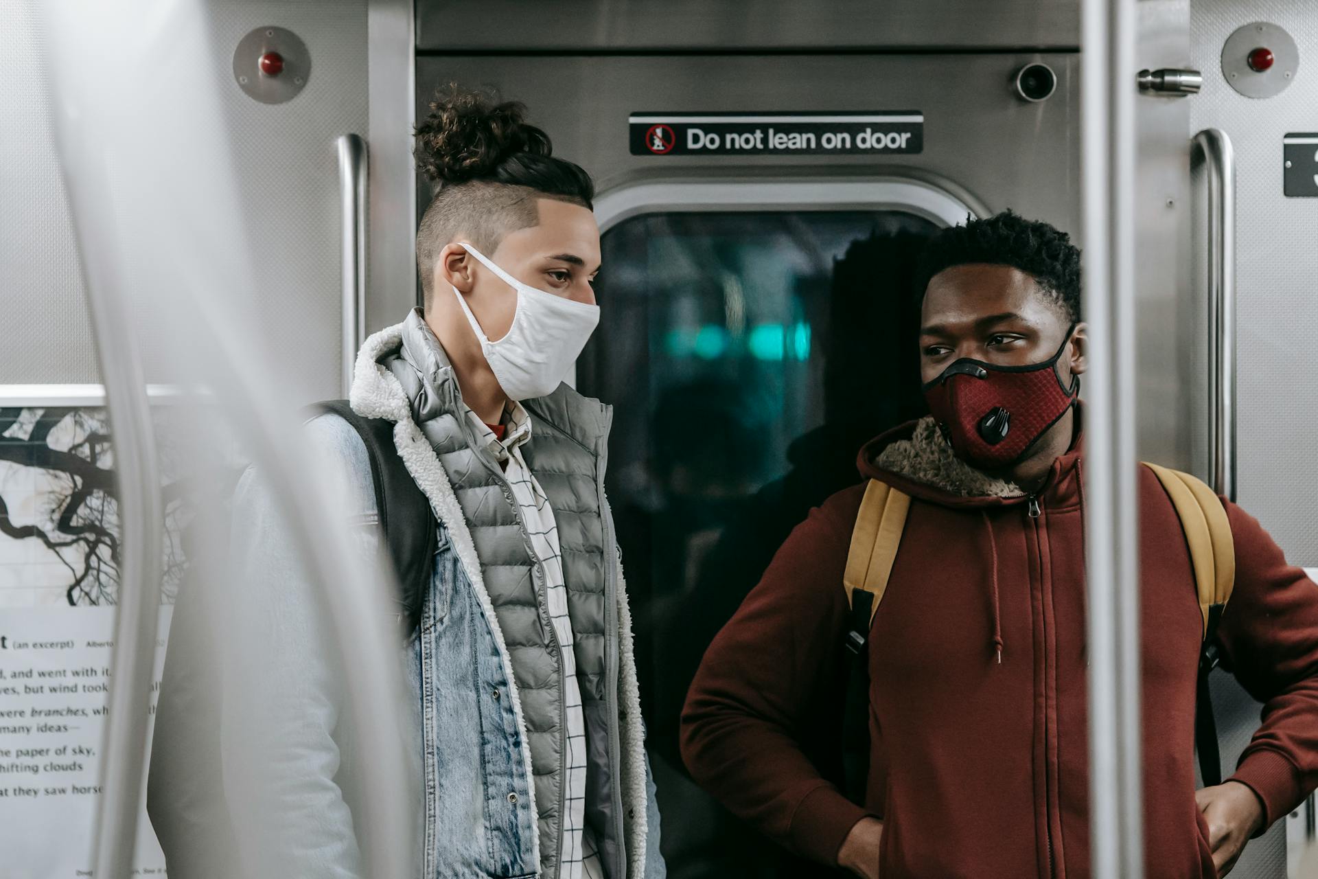 Two young men wearing protective masks converse on a subway train