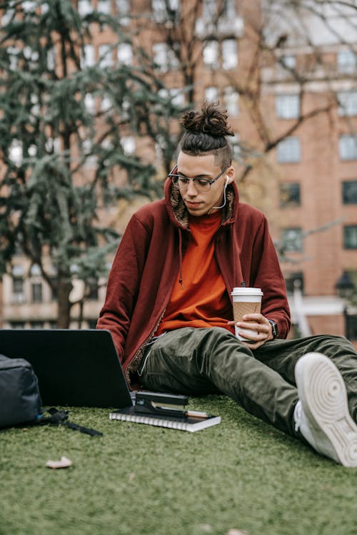 Young man with takeaway hot drink browsing internet on portable computer while studying in city park