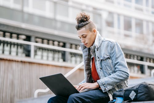 Young focused man in casual wear with modern haircut surfing internet on netbook while studying on urban staircase