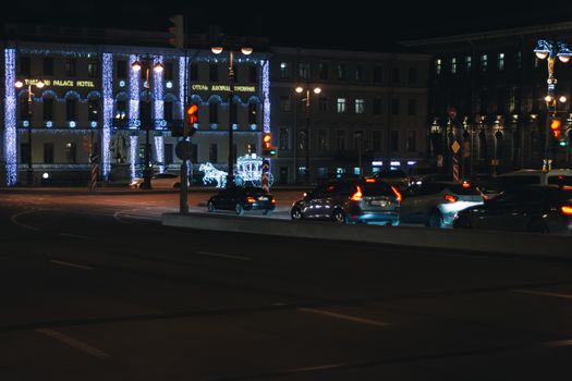 Modern cars driving on asphalt road near historic building decorated with glowing garlands at night in Saint Petersburg