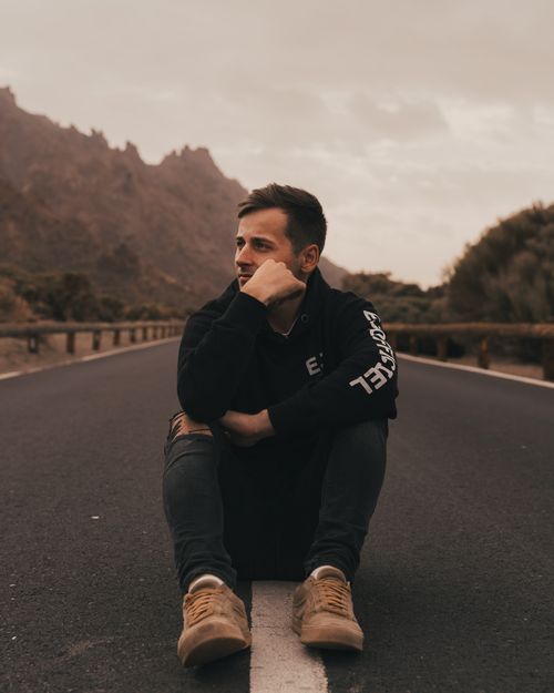 Free A contemplative young man sits on a road surrounded by a mountainous landscape, evoking a sense of adventure and solitude. Stock Photo