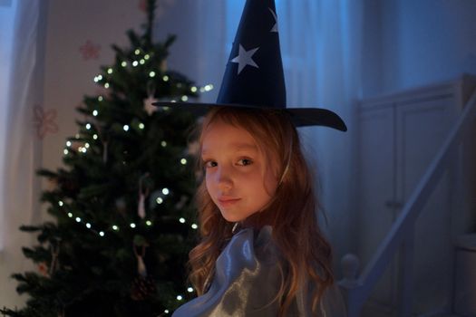 A young girl with blonde hair in a witch hat stands by a festive Christmas tree.