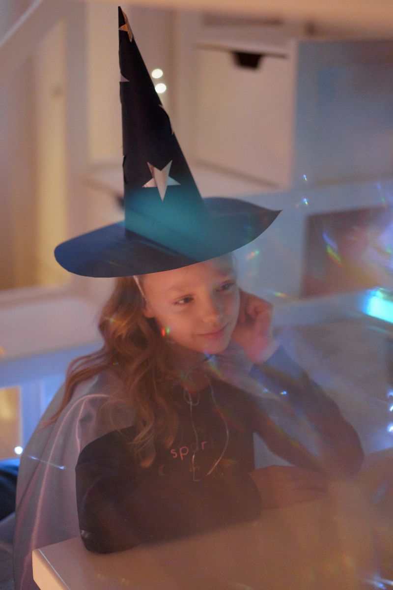 A young girl wearing a black wizard hat and white cloak holding a wand