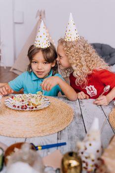 Two kids in party hats enjoying cake at a joyful indoor birthday celebration.
