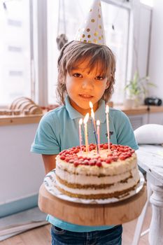Young boy in party hat holding a birthday cake with lit candles indoors.