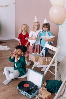 A group of children enjoy a birthday party indoors with gifts, balloons, and music.