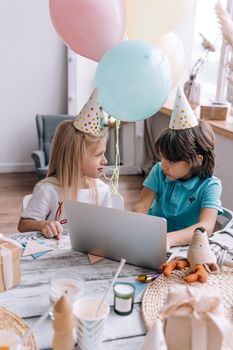 Two children enjoying a virtual birthday party indoors with balloons and a laptop.
