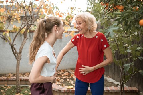 Free Two women laughing and talking under fruit trees in a garden, enjoying a sunny day. Stock Photo