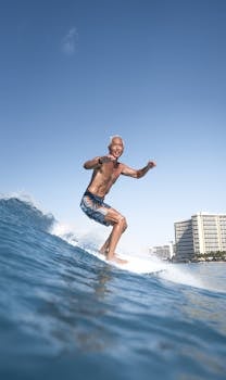 A senior man surfing on ocean waves with buildings in the background under a clear blue sky.