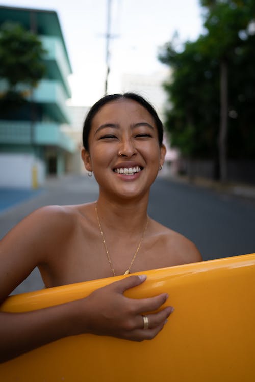 Smiling ethnic lady standing with surfboard in town
