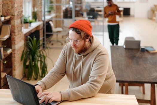 A bearded man in a hoodie works on a laptop in a modern, casual office setting.