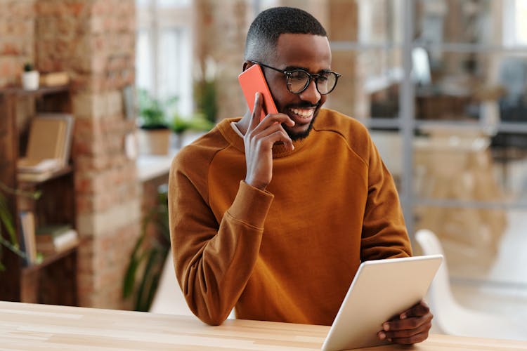 A Man In Brown Sweater Talking On The Phone While Holding His Tablet