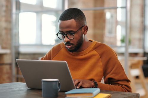Free Young man with glasses works on laptop at a wooden table indoors. Warm, productive atmosphere. Stock Photo